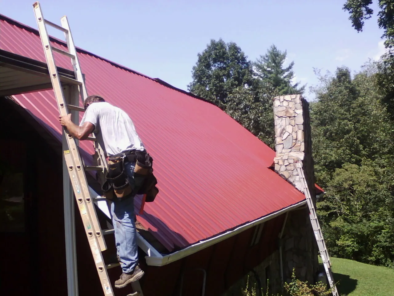Roofer on a ladder beside a red metal roof and stone chimney — Soffit & Fascia Repair by Roofing Brothers in Dalbo